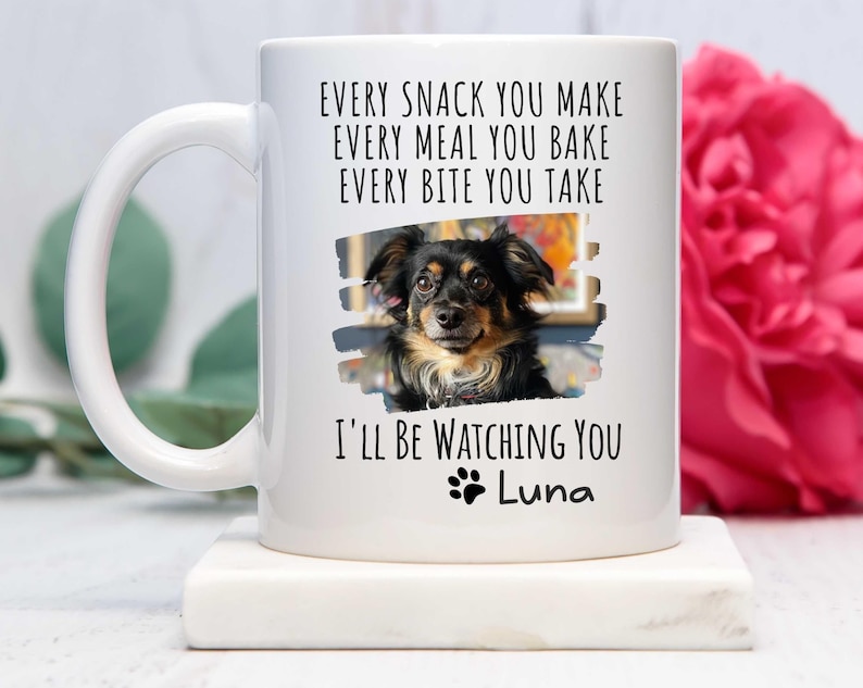 May include: White ceramic coffee mug with a black paw print and the text "EVERY SNACK YOU MAKE EVERY MEAL YOU BAKE EVERY BITE YOU TAKE I'LL BE WATCHING YOU Luna". A photo of a brown and black dog is in the center of the mug.