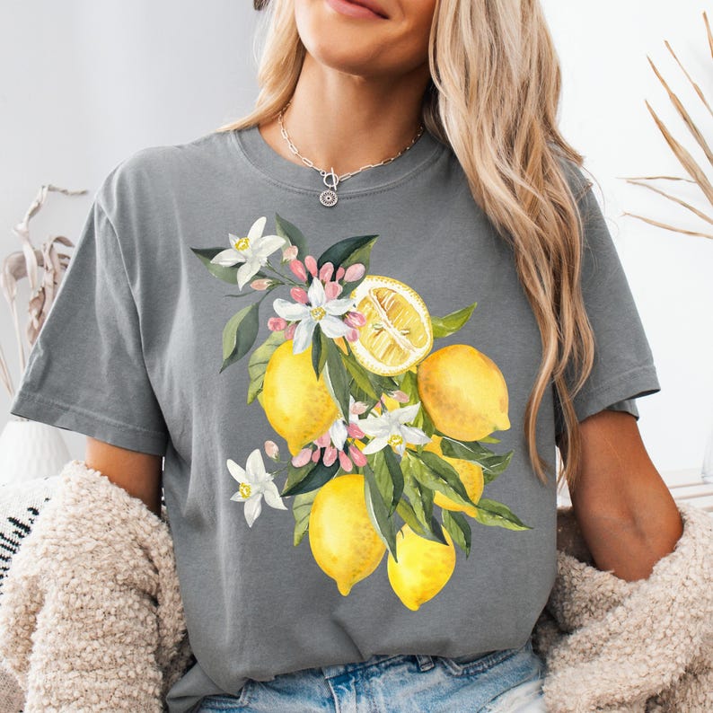 May include: Gray t-shirt featuring a watercolor design of lemons, white blossoms, and green leaves. The lemons are yellow, and one is sliced to reveal the inside. The shirt is a comfortable, casual style.