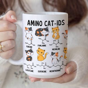Może przedstawiać: Biały kubek ceramiczny z kolorową ilustracją kot&oacute;w i struktur chemicznych. Na kubku nadrukowany jest tekst "AMINO CAT-IDS". Struktury chemiczne są oznaczone nazwami takimi jak "FURCINE", "PAWLANINE", "MEOWLINE", "LUCATLINE", "ISOFURCINE" i "MEOWTHIONINE".