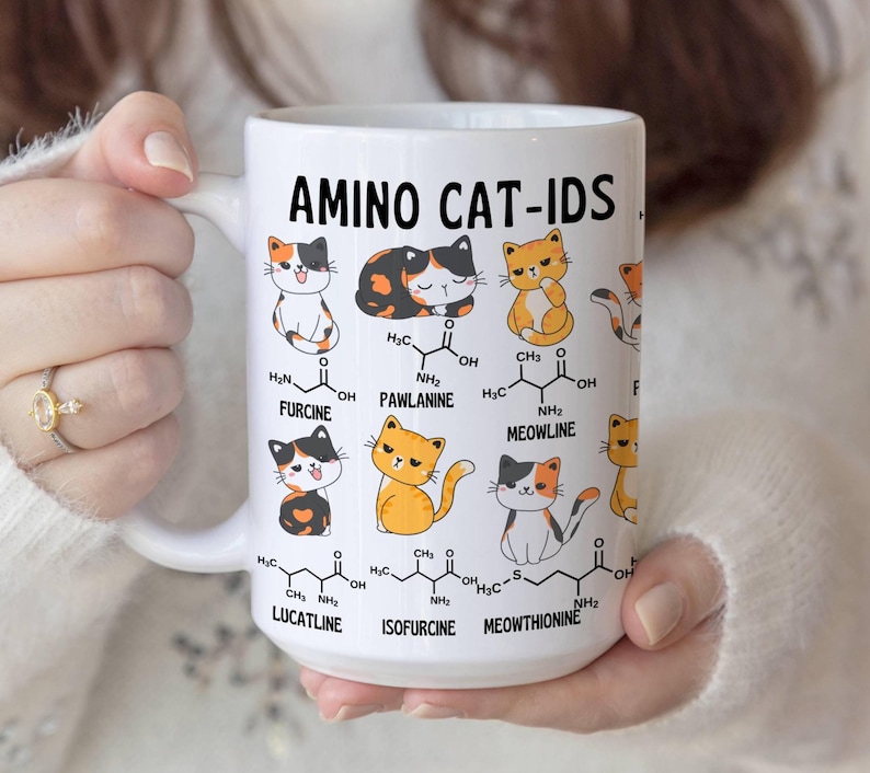 Może przedstawiać: Biały kubek ceramiczny z kolorową ilustracją kot&oacute;w i struktur chemicznych. Na kubku nadrukowany jest tekst "AMINO CAT-IDS". Struktury chemiczne są oznaczone nazwami takimi jak "FURCINE", "PAWLANINE", "MEOWLINE", "LUCATLINE", "ISOFURCINE" i "MEOWTHIONINE".