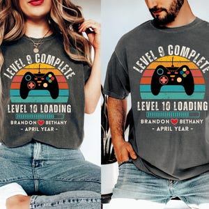 May include: Two gray t-shirts with a retro video game controller graphic and the text "Level 9 Complete" and "Level 10 Loading". The shirts also include the text "Brandon Bethany - April Year - "