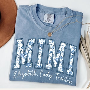 May include: A light blue t-shirt with a white floral design spelling out "MIMI" in large letters. Below the floral design is the text "Elizabeth. Cody. Trenton".