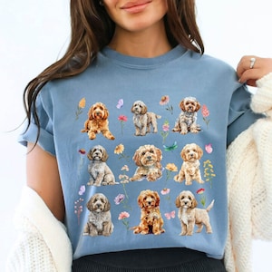 Peut inclure: T-shirt bleu clair orné d'un motif de Cockapoos, chiens adorables, entourés de fleurs et de papillons colorés. Les chiens sont représentés dans diverses poses, créant un design charmant.