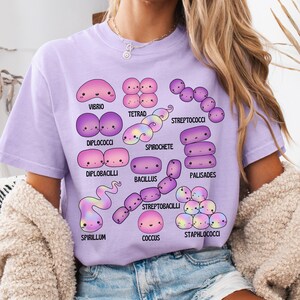 May include: A lilac t-shirt displaying cartoon bacteria illustrations, such as Vibrio, Tetrad, and Spirochete. Each bacterium is uniquely shaped and labelled with its name in black text. The t-shirt is a casual style.