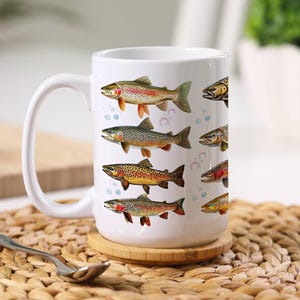 May include: White ceramic coffee mug featuring colorful illustrations of various fish species. The mug has a large handle and is decorated with images of trout, with bubbles in the background. The mug is sitting on a wooden coaster.