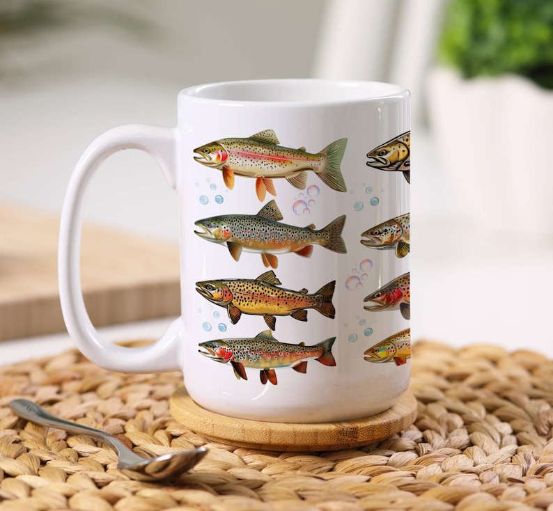 May include: White ceramic coffee mug featuring colorful illustrations of various fish species. The mug has a large handle and is decorated with images of trout, with bubbles in the background. The mug is sitting on a wooden coaster.