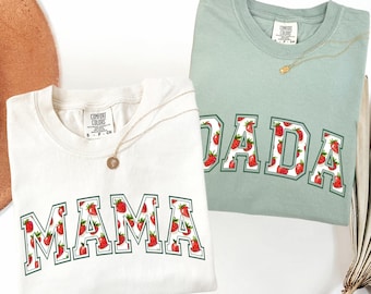 May include: Two Comfort Colors crewneck t-shirts, one cream and one sage green. The cream shirt says "MAMA" and the green shirt says "DADA", both in a collegiate font filled with strawberries. Each shirt has a gold necklace.