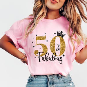 May include: A light pink short-sleeve t-shirt featuring a graphic design with the gold glitter numbers 50, a black crown icon, and the black script text & Fabulous. The design includes small black star accents surrounding the central text.