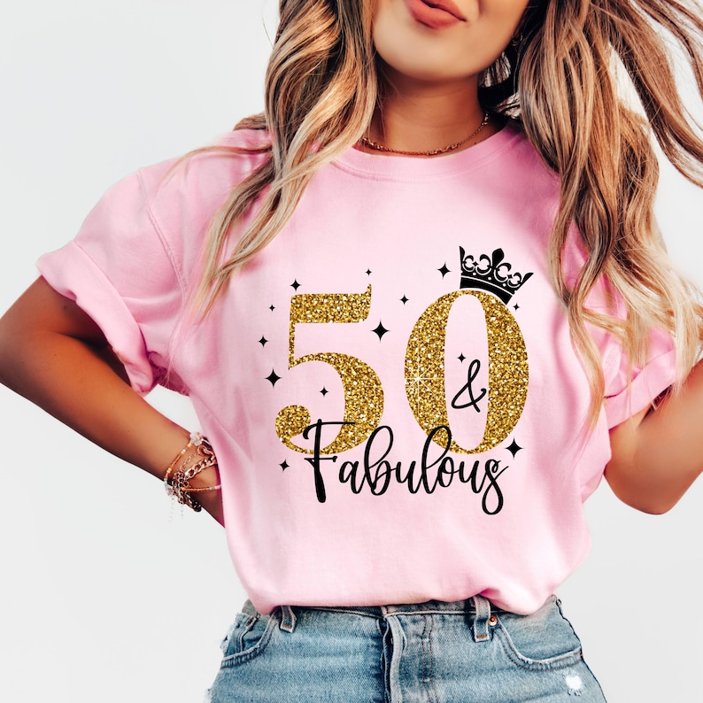 May include: A light pink short-sleeve t-shirt featuring a graphic design with the gold glitter numbers 50, a black crown icon, and the black script text & Fabulous. The design includes small black star accents surrounding the central text.