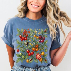 Op de afbeelding: Blauw T-shirt met een kleurrijk tomatenplantontwerp. De afbeelding toont rode, oranje en groene tomaten, gele bloemen, een hommel en lieveheersbeestjes. Het shirt heeft een casual stijl.