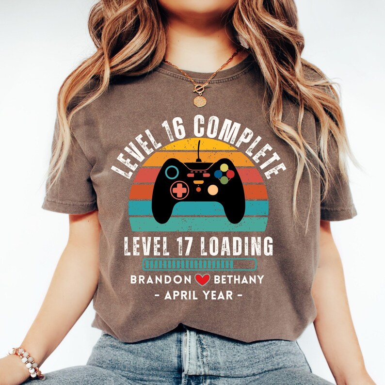 May include: A brown t-shirt with a retro-style graphic of a video game controller and the text "Level 16 Complete Level 17 Loading". The graphic also includes a progress bar with the names "Brandon" and "Bethany" and the text "April Year".
