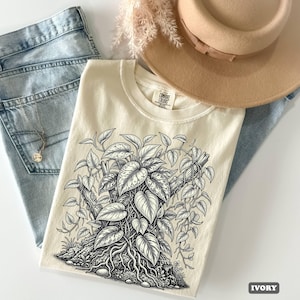 以下が含まれることがあります： アイボリー色のTシャツで、質感のある丸太の上で這うように成長するポトスの植物の白黒線画イラストが描かれています。TシャツにはクルーネックとComfort Colorsのブランドラベルがあり、詳細なハート型の葉のデザインが特徴です。