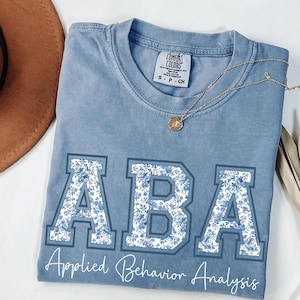 以下が含まれることがあります： 花柄の「ABA」の文字が入った青色のコンフォートカラーズTシャツ。「Applied Behavior Analysis」の文字が文字の下にプリントされています。ペンダント付きのゴールドネックレスがTシャツの上にかけられています。