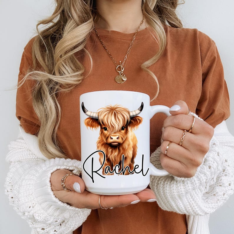 Może przedstawiać: Biały kubek ceramiczny z ilustracją brązowej krowy szkockiej g&oacute;rskiej i imieniem "Rachel" napisanym czarnym, ozdobnym pismem.