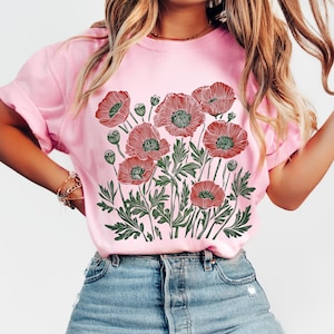 May include: Pink t-shirt featuring a detailed illustration of red poppies with green leaves and buds. The floral design is centered on the shirt. The shirt is made of a soft material and has rolled-up sleeves.