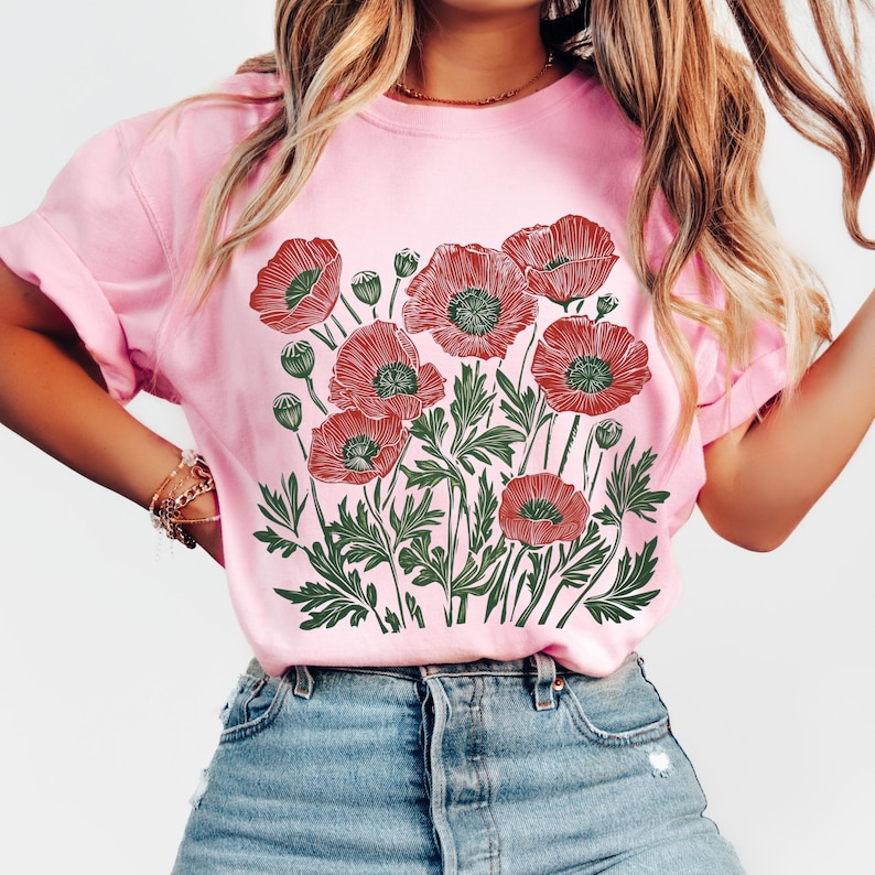 May include: Pink t-shirt featuring a detailed illustration of red poppies with green leaves and buds. The floral design is centered on the shirt. The shirt is made of a soft material and has rolled-up sleeves.