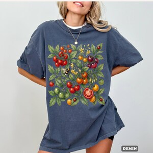 Op de afbeelding: Donkerblauw T-shirt met een kleurrijke illustratie van tomaten, bladeren, bloemen, een bij en lieveheersbeestjes. Het ontwerp bevat rode, groene, gele en oranje tomaten. Het woord "DEMIN" staat rechtsonder in het wit gedrukt.