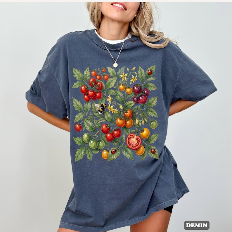 Op de afbeelding: Donkerblauw T-shirt met een kleurrijke illustratie van tomaten, bladeren, bloemen, een bij en lieveheersbeestjes. Het ontwerp bevat rode, groene, gele en oranje tomaten. Het woord "DEMIN" staat rechtsonder in het wit gedrukt.