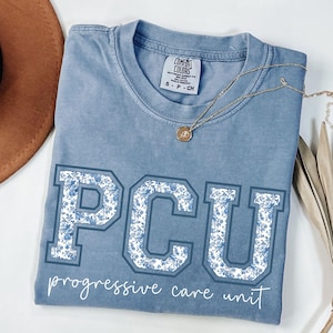 May include: A blue Comfort Colors t-shirt with the letters "PCU" in a floral design. The words "progressive care unit" are printed below the letters. A gold necklace with a pendant is draped across the shirt.