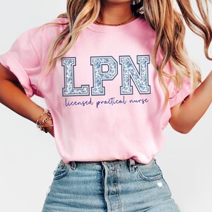 May include: A light pink t-shirt with the letters "LPN" in a blue floral pattern. Below the letters, the words "licensed practical nurse" are written in a dark purple script. The shirt is made of a soft material.