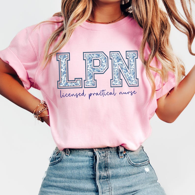 May include: A light pink t-shirt with the letters "LPN" in a blue floral pattern. Below the letters, the words "licensed practical nurse" are written in a dark purple script. The shirt is made of a soft material.