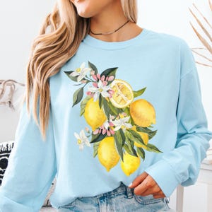 May include: A light blue sweatshirt featuring a watercolor illustration of lemons, blossoms, and green leaves. The design includes whole and sliced lemons, with white and pink flowers. The long-sleeved top is made of a soft material.