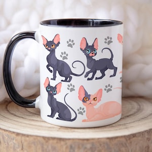 Puede incluir: Taza de cerámica blanca con borde y asa negros. La taza presenta un patrón repetido de gatos sin pelo negros y rosas con huellas de patas.