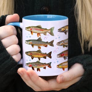 May include: White ceramic mug with a blue interior and handle, featuring illustrations of various colorful fish. The fish designs include brown, yellow, red, and green hues. The mug is held by a person.