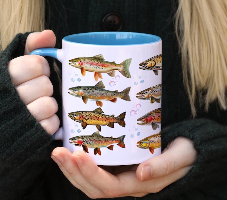 May include: White ceramic mug with a blue interior and handle, featuring illustrations of various colorful fish. The fish designs include brown, yellow, red, and green hues. The mug is held by a person.