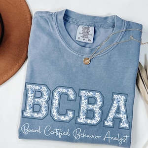 Pode incluir: Uma camiseta Comfort Colors azul claro com as letras "BCBA" em um design floral. O texto "Board Certified Behavior Analyst" está abaixo. Um colar de ouro com um pingente está na camiseta.