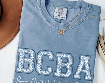 May include: A light blue Comfort Colors t-shirt with the letters "BCBA" in a floral design. The text "Board Certified Behavior Analyst" is below the letters. A gold necklace with a pendant is on the shirt.