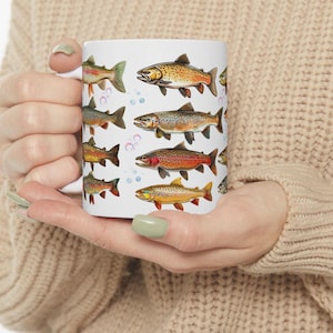 May include: White ceramic mug featuring colorful illustrations of various fish species. The fish are depicted in shades of orange, brown, and green, with bubble accents. The mug is held in a person's hands, showcasing the design.