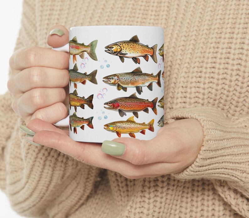 May include: White ceramic mug featuring colorful illustrations of various fish species. The fish are depicted in shades of orange, brown, and green, with bubble accents. The mug is held in a person's hands, showcasing the design.