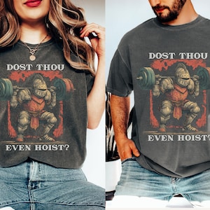 Pode incluir: Duas camisetas cinza escuro com o texto "DOST THOU EVEN HOIST?" acima e abaixo de um gráfico de um cavaleiro levantando pesos. O cavaleiro está de armadura completa, agachado com uma barra. As camisas são usadas por duas pessoas.
