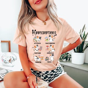 Könnte beinhalten: Ein pfirsichfarbenes T-Shirt mit dem Text "Purrscriptions" und Illustrationen von Katzen mit verschiedenen Medikamenten. Das Shirt hat ein verspieltes Design mit katzenbezogenen Wortspielen über gängige Medikamente, wie "Kittyprofen" und "Purrzac".