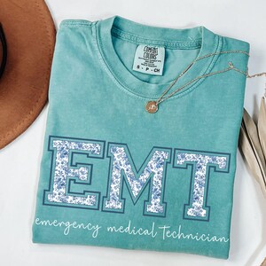 Puede incluir: Camiseta Comfort Colors de color turquesa con la palabra "EMT" en letras grandes y en bloque rellenas con un estampado floral azul. Las palabras "emergency medical technician" est&aacute;n impresas debajo. Un collar dorado con un colgante est&aacute; sobre la camisa.