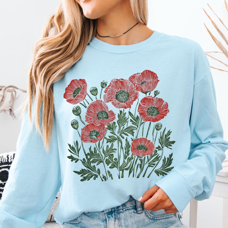 May include: A light blue sweatshirt featuring a red poppy floral design. The graphic includes detailed red poppies with green centers and leaves. The long-sleeved top is a casual style.