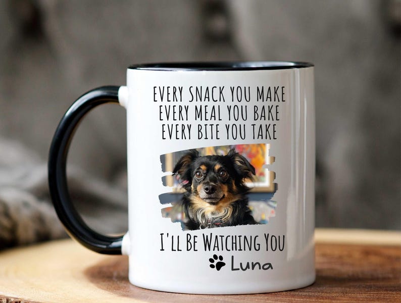May include: White ceramic mug with a black rim and handle. The mug features a black and white photo of a dog with the text "EVERY SNACK YOU MAKE EVERY MEAL YOU BAKE EVERY BITE YOU TAKE I'LL BE WATCHING YOU Luna".