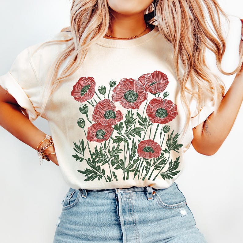 May include: Cream-colored t-shirt featuring a red and green floral design of poppies. The graphic includes detailed line art of the flowers, leaves, and buds. The shirt is made of a soft material and has short sleeves.