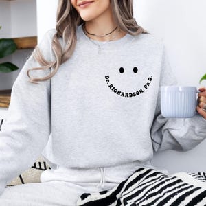 May include: A light gray sweatshirt with a smiley face design. The smiley face includes two black dots for eyes and a curved line with the text "Dr. Richardson, Ph.D." The person is holding a light blue mug.