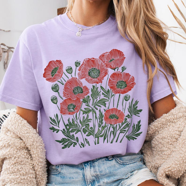 May include: A light purple t-shirt featuring a graphic of red poppies with green leaves and stems. The floral design is centered on the shirt. The shirt has short sleeves and a crew neck.