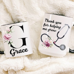 Puede incluir: Dos tazas de cerámica blanca con asas negras. Una taza presenta el nombre "Grace" con un diseño floral de "G". La otra taza tiene un gráfico de estetoscopio y el texto "Thank you for helping me grow."