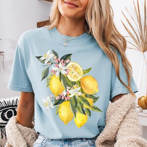 May include: Light blue t-shirt featuring a watercolor illustration of lemons, blossoms, and green leaves. The design includes several yellow lemons, white flowers, and pink accents. The shirt is a comfortable, casual style.