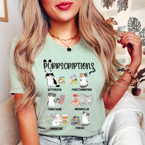 Könnte beinhalten: Hellgrünes T-Shirt mit dem Text "Purrscriptions" und Illustrationen von Katzen und Medikamentenflaschen. Das Shirt zeigt verschiedene katzenbezogene Wortspiele auf Medikamentennamen wie "Kittyprofen" und "Purrzac".