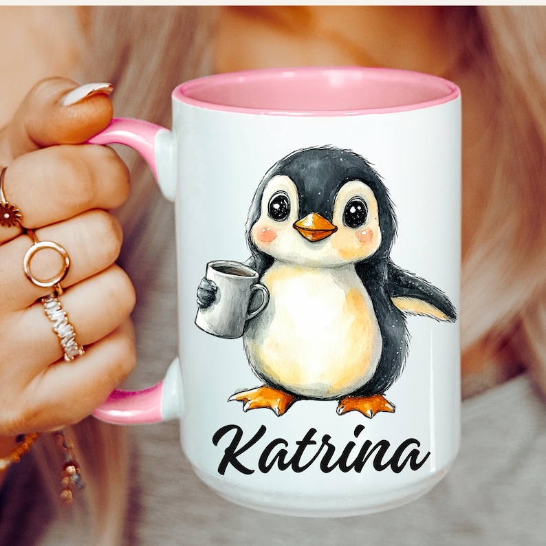 May include: White ceramic mug with a pink interior and handle. The mug features a watercolor illustration of a cartoon penguin holding a coffee cup. The penguin has black and gray feathers, an orange beak and feet, and the name "Katrina" in black script.