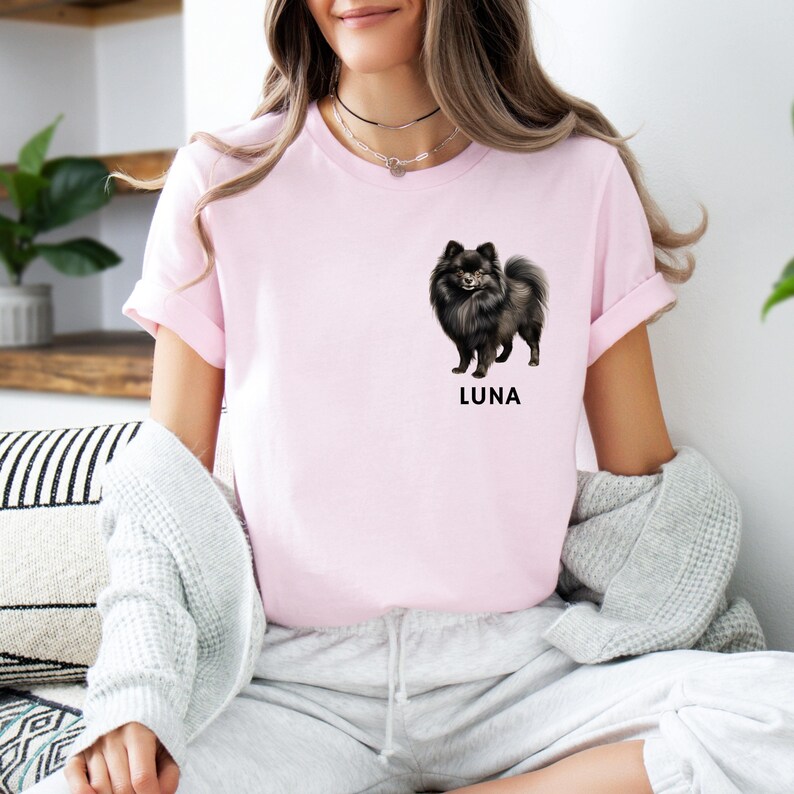 May include: A pink t-shirt with a black and white illustration of a dog on the front. The dog is a Pomeranian and the text "LUNA" is printed below the dog.