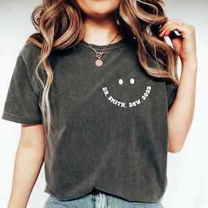 Könnte beinhalten: Dunkelgraues T-Shirt mit einem Smiley-Gesicht-Design und dem Text "DR. SMITH, DSW 2025" in Weiß. Das Shirt hat einen Rundhalsausschnitt und kurze Ärmel. Der Stoff wirkt weich und leicht verwaschen.
