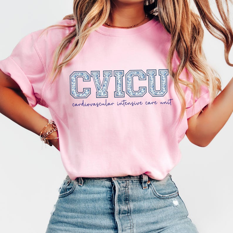 May include: A light pink t-shirt with the text "CVICU" in a blue floral design. Below the letters, the words "cardiovascular intensive care unit" are written in a smaller, purple font. The shirt is made of a soft material.