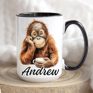 May include: White ceramic mug with a black interior and handle. The mug features a watercolor illustration of an orangutan holding a cup, with the name "Andrew" in black script. The mug is sitting on a wooden surface.