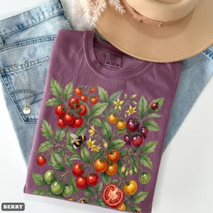 Op de afbeelding: Een bessenkleurig T-shirt met een gedetailleerde illustratie van een tomatenplant met rode, oranje en groene tomaten, gele bloemen, een hommel en een lieveheersbeestje. Het shirt is opgevouwen op een spijkerbroek, met een ketting en een beige hoed.